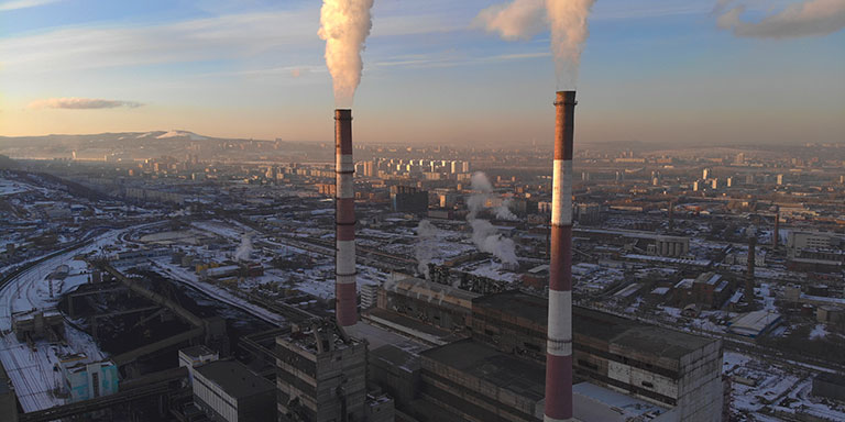 Two tall industrial chimneys release gas into the air in a large city