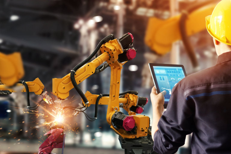 Sparks fly from the work of three yellow robotic arms as a worker monitors a tablet