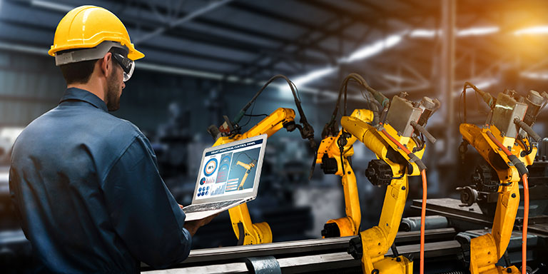 A worker wearing a hard hat monitors a laptop in front of yellow robotic arms