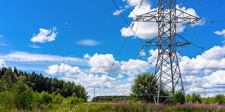 An electrical transmission tower stands in a field against a bright blue sky and white clouds